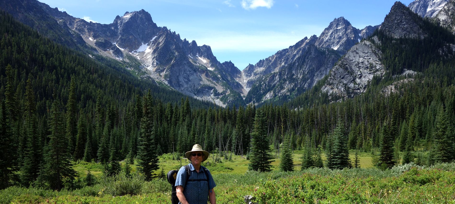 Summer view of Innkeeper hiking in lush green meadows framed by jagged mountain peaks.